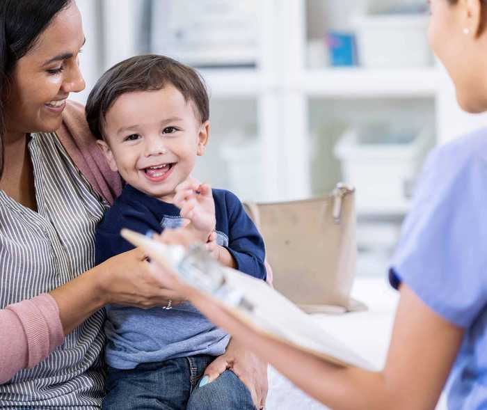 Kleiner Junge sitzt auf dem Schoß seiner Mutter gegenüber von einer Arzthelferin und freut sich 