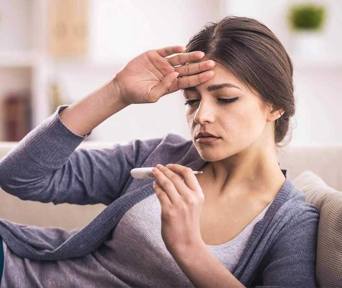 Frau fasst sich an die Stirn und schaut auf ein Fieberthermometer