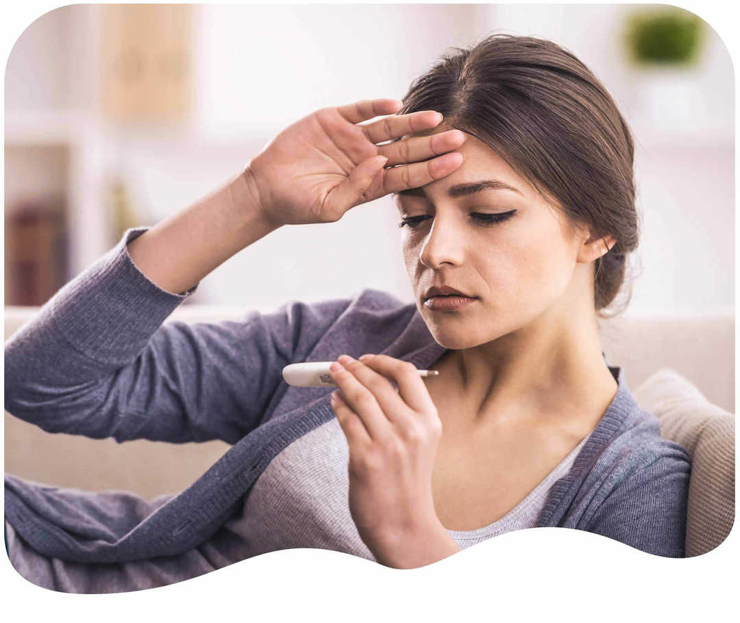 Frau fasst sich an die Stirn und schaut auf ein Fieberthermometer