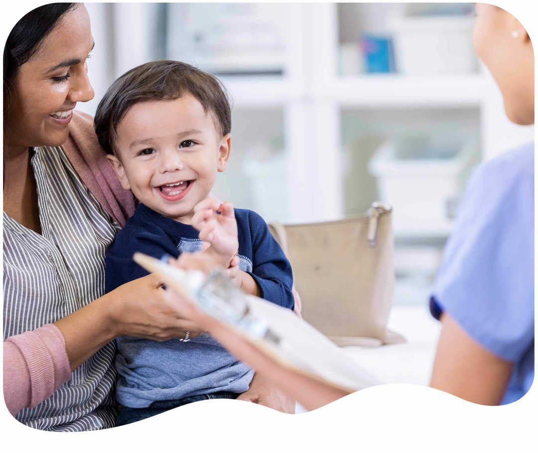 Kleiner Junge sitzt auf dem Schoß seiner Mutter gegenüber von einer Arzthelferin und freut sich 